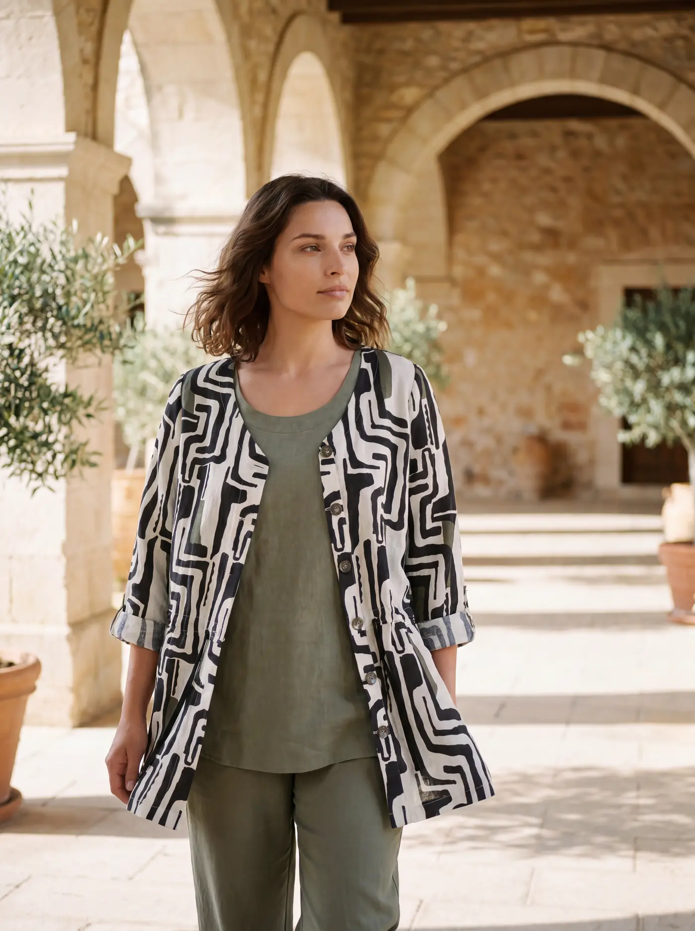 Curvy-KI-Model trägt die Longbluse offen wie ein Jacken-Layering über dem olivgrünen Leinen-Co-ord in einem Kreuzgang